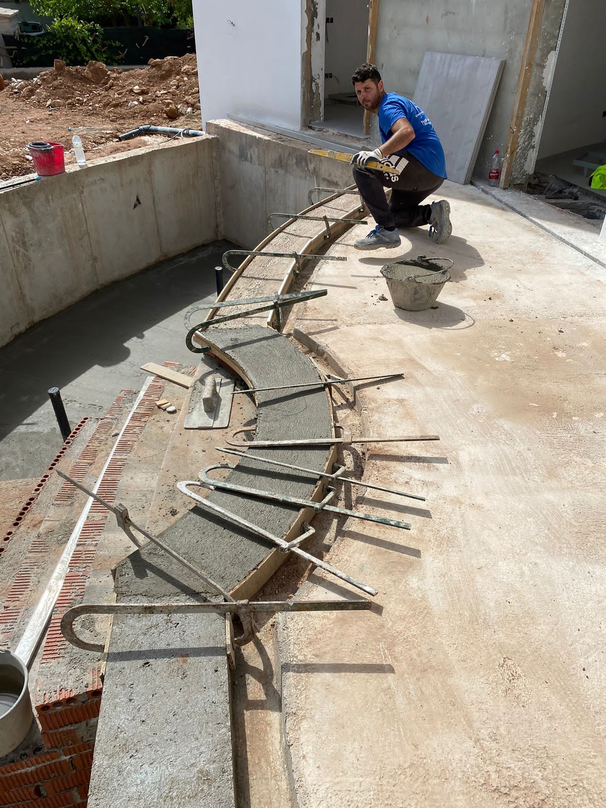 Uno del equipo, en cuclillas, junto al encofrado de la escalera curva de una piscina, con los hierros puestos y el cubo de mortero al lado