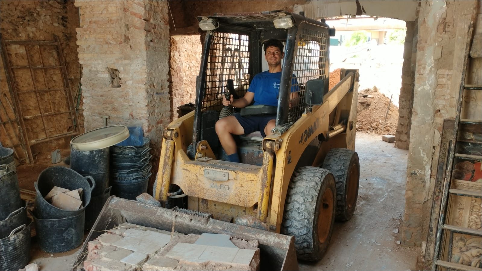 Uno del equipo a los mandos de una minicargadora con escombro en la pala, en una casa en rehabilitación con los muros de ladrillo a la vista
