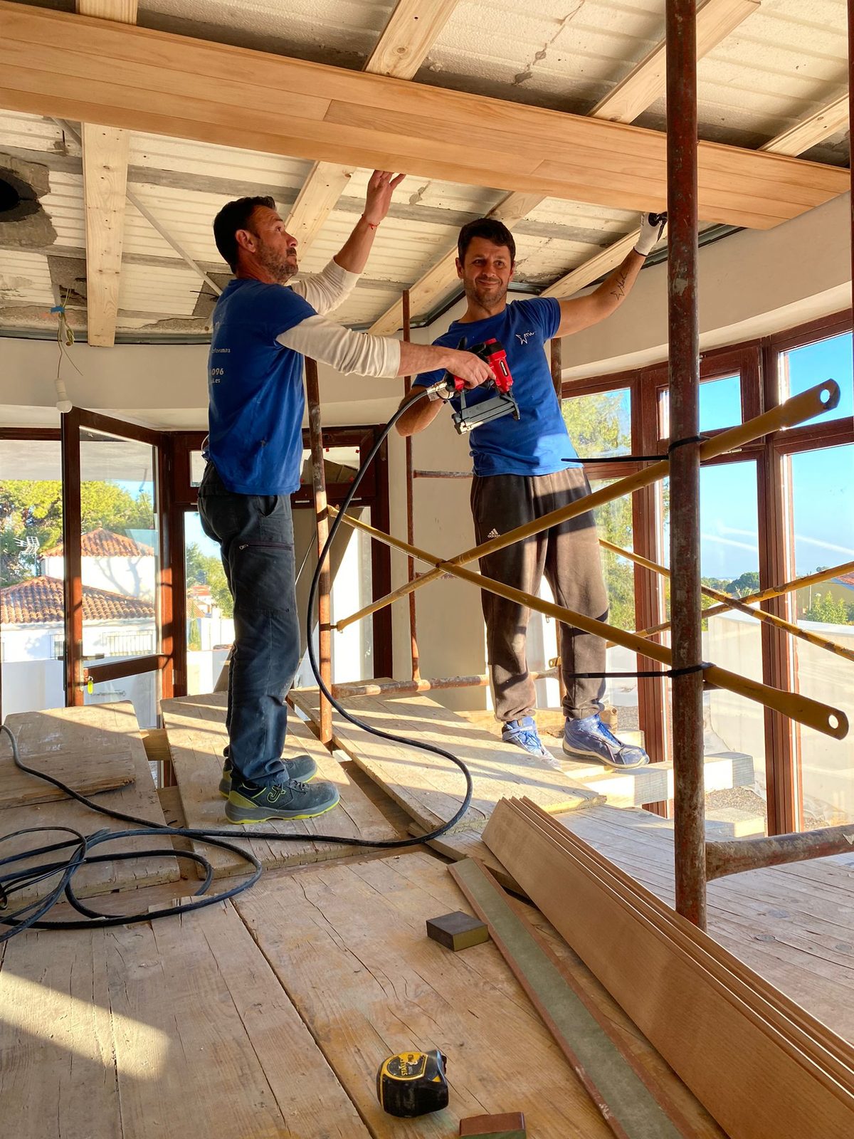 Dos del equipo fijan con una clavadora una tabla de madera al techo de una vivienda con grandes ventanales